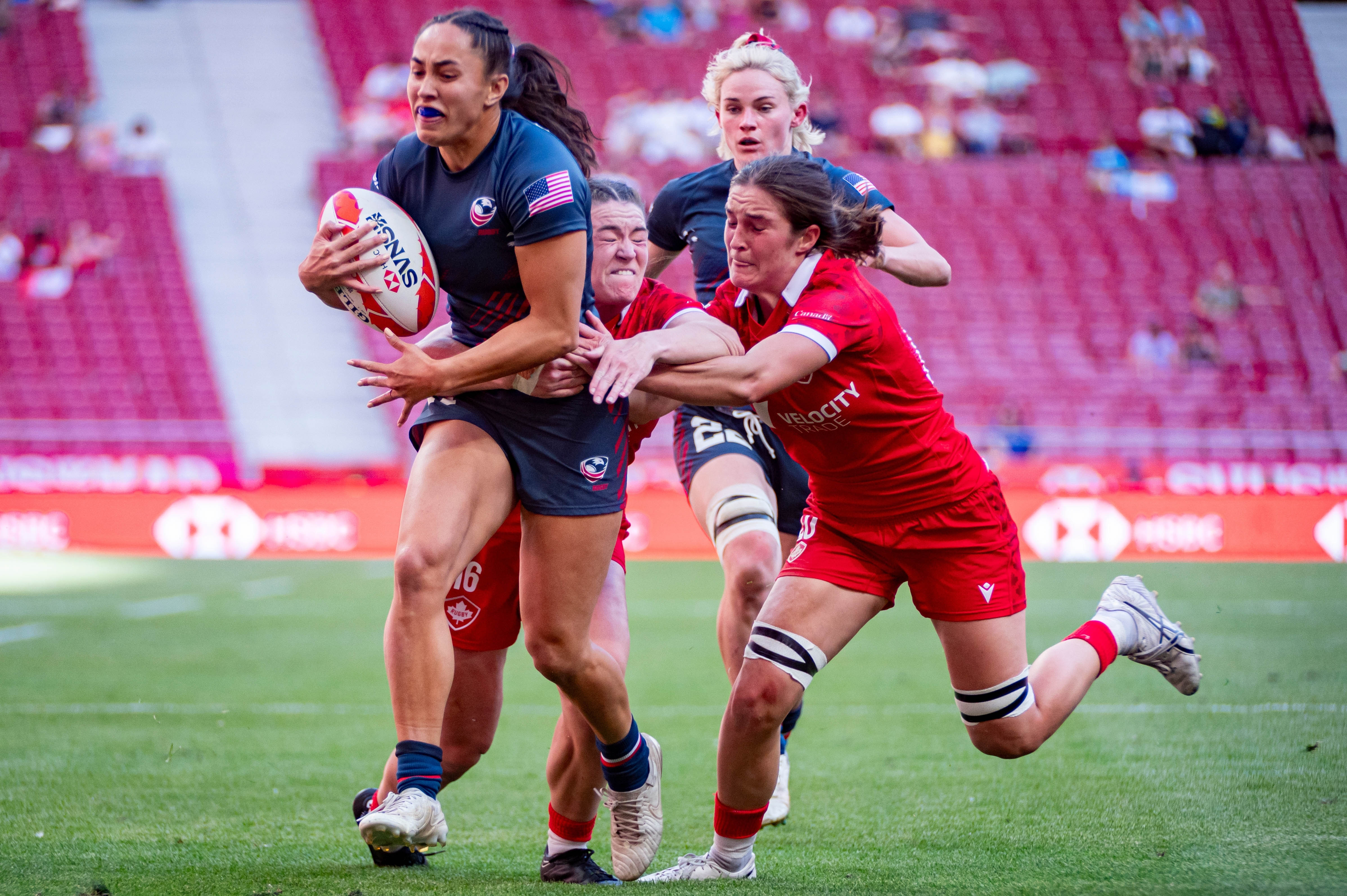 Rugby game at the Olympics - four women running, one with the ball, two attempting to grab her for a tackle, and a fourth chasing behind. Woman with the ball is an American athlete. Rugby game at the Olympics - four women running, one with the ball, two attempting to grab her for a tackle, and a fourth chasing behind. Woman with the ball is an American athlete.